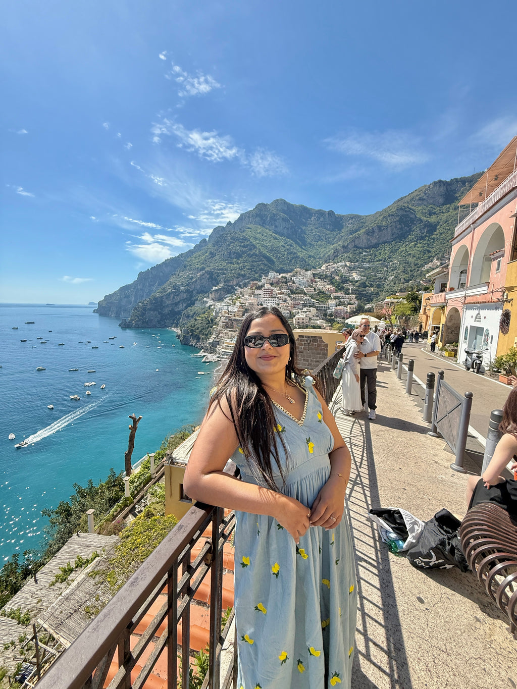 Woman in a light blue dress with yellow patterns standing on a scenic coastal path with mountains and sea in the background.